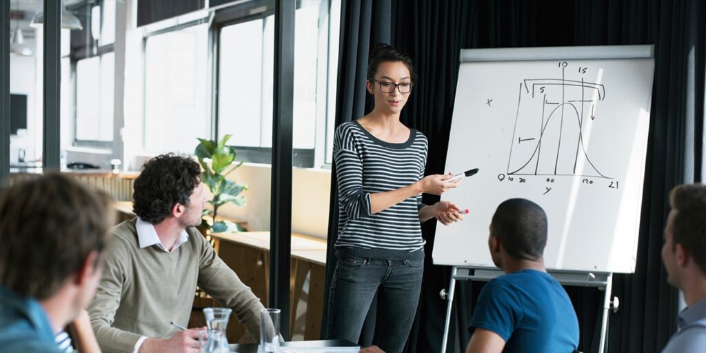 a woman stands at a whiteboard at the front of a meeting room presenting a formula