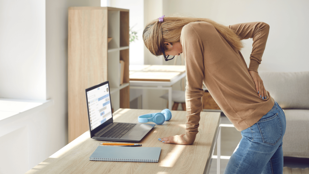 a pained woman leans on her desk with a hand on her lower back